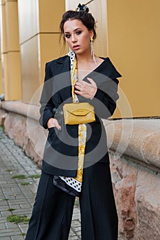Young stylish beautiful woman fashion model is posing in street, wearing pantsuit, having purse on her waist