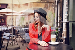 Young stylish beautiful woman sitting in city cafe in red jacket, street style, drinking aromatic coffee. Elegant girl in hat