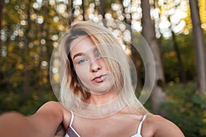 Young stylish blonde woman in a striped pink fashionable sundress makes selfie standing in the forest on a bright sunny summer day