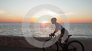 Young stylish brunette woman rides bike on coastline at sunrise. Brunette girl in helmet glasses and stylish clothes is pedaling o