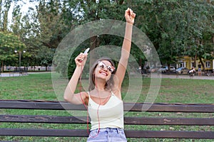 Young stylish cute girl is listening to music in headphones on mobile phone and dancing in the park. Pretty woman is relaxing and