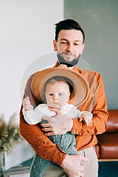 A young stylish father in trend orange shirt plays with his young son in a brown hat in his arms in a spacious stylish