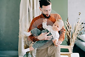 A young stylish father in trend orange shirt plays with his young son in a brown hat in his arms in a spacious stylish