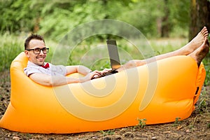 Young stylish freelancer man in glasses working at laptop sitting on Inflatable mattress in garden. Online education