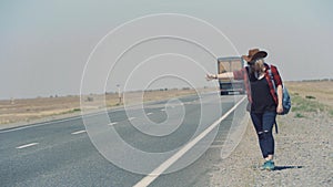 Young stylish girl in cowboy clothes doing hitch-hiking in desert, on the road. She carries backpack and hat. Pass