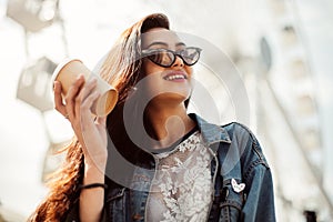 Young stylish girl enjoys summer and vacation. Brunette in trendy sunglasses holding coffee to go