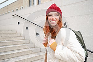 Young stylish girl traveller goes sightseeing, travels around city with backpack, tourist smiles and looks around