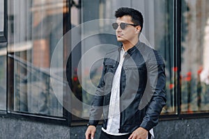 Young stylish guy in glasses in black leather jacket on glass background