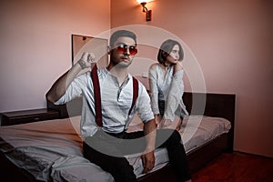 A young stylish guy in glasses and a young girl together sitting on the bed. the guy closer to the girl further. selective focus