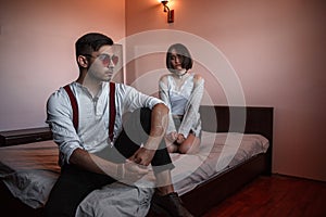 A young stylish guy in glasses and a young girl together sitting on the bed. the guy closer to the girl further. selective focus