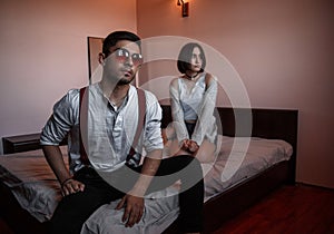 A young stylish guy in glasses and a young girl together sitting on the bed. the guy closer to the girl further. selective focus