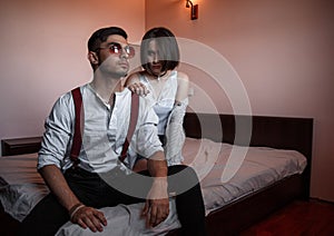 A young stylish guy in glasses and a young girl together sitting on the bed. the guy closer to the girl further. selective focus