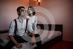 A young stylish guy in glasses and a young girl together sitting on the bed. the guy closer to the girl further. selective focus