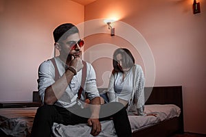 A young stylish guy in glasses and a young girl together sitting on the bed. the guy closer to the girl further. selective focus