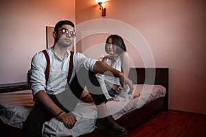 A young stylish guy in glasses and a young girl together sitting on the bed. the guy closer to the girl further. selective focus