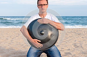 Young stylish guy sitting on the sand beach and holding a handpan or hang with sea On Background. The Hang is