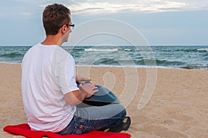 Young stylish guy sitting on the sand beach and playing handpan or hang with sea On Background. The Hang is traditional