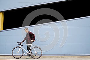 Young stylish guy walks with a white bicycle in front of a blue wall, a student with a backpack returns