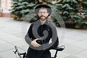 Young stylish handsome man in glasses and hat standing with his bicycle on the street