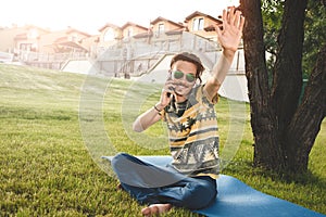 Young stylish handsome smiling man with sunglasses is sitting on grass, resting and talking on the phone. waves his hand welcoming