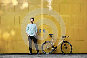 A young stylish hipster caucasian man posing next to his bicycle on the yellow background.