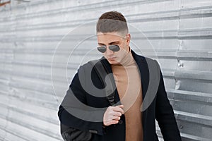 Young stylish hipster man in black sunglasses with a stylish hairstyle in a beige knitted sweater with a backpack in a trendy coat