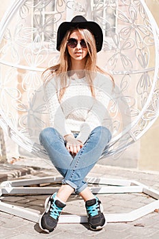 Young stylish hipster woman in sunglasses in fashionable spring clothes is sitting on a metal vintage chair on a sunny warm day.