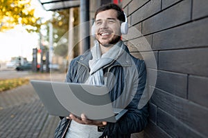 young stylish man listening to music in headphones with enjoyment covering his eyes with a laptop in his hands outside