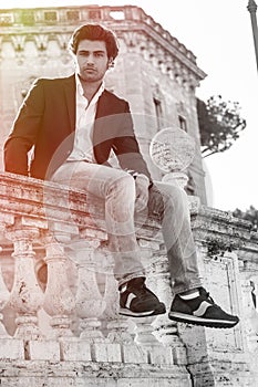Young stylish man model sitting on old historic low wall. Rome, Italy