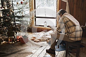 Young stylish man is stoking stove, reading cook. Celebrating new year eve in cozy wooden country house in winter forest