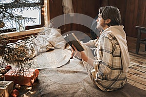 Young stylish man is stoking stove, reading cook. Celebrating new year eve in cozy wooden country house in winter forest