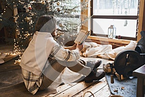 Young stylish man is stoking stove, reading cook. Celebrating new year eve in cozy wooden country house in winter forest