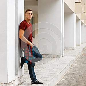 Young stylish man in stylish red t-shirt in a plaid trendy shirt in fashionable blue jeans in sneakers posing outdoors near a