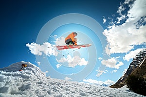 A young stylish man in sunglasses and a cap performs a trick in jumping with a kicker of snow against the blue sky and