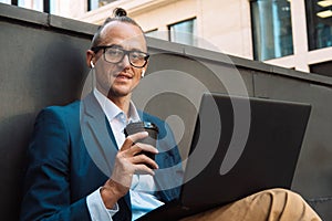 Young stylish man working on laptop computer while sitting on bench outdoors and drinking coffee.