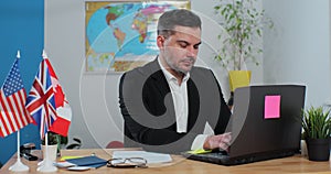 Young and stylish man works in a travel agency, on laptop computer, sitting at a table on the background of a world map