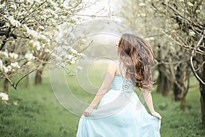 A young stylish model girl in a beautiful fashionable long green dress stands in a summer park near lush flower bushes
