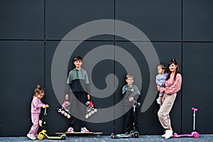 Young stylish mother with four kids outdoor against black modern wall. Sports family spend free time outdoors with scooters and