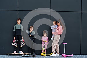 Young stylish mother with four kids outdoor against black modern wall. Sports family spend free time outdoors with scooters and