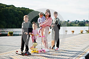 Young stylish mother with four kids outdoor. Sports family spend free time outdoors with scooters and skates