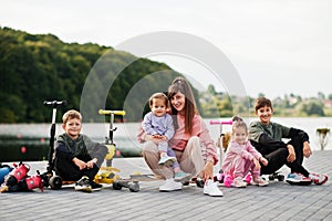 Young stylish mother with four kids outdoor. Sports family spend free time outdoors with scooters and skates