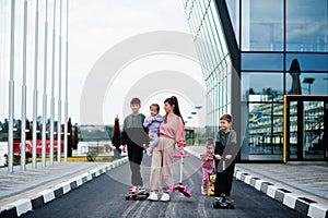 Young stylish mother with four kids outdoor. Sports family spend free time outdoors with scooters and skates