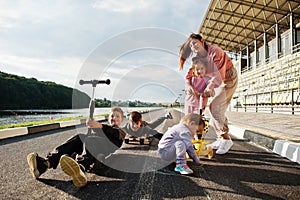 Young stylish mother with four kids outdoor. Sports family spend free time outdoors with scooters and skates