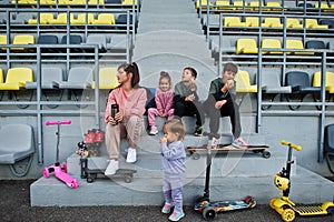 Young stylish mother with four kids sitting on the sports podium at the stadium, eat apple and drink water. Family spend free time