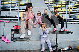 Young stylish mother with four kids sitting on the sports podium at the stadium, eat apple and drink water. Family spend free time
