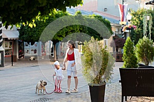 A young stylish mother in white leggings and a white jacket is walking in the city with her little daughter and a dog