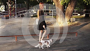 Young, stylish red headed girl walking by empty skatepark with her loving dog. Slow motion of pretty girl in black dress