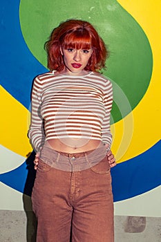 Young stylish redhead Caucasian woman in retro style of 70s posing at wall looking into camera