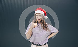 Young stylish trendy woman isolated over grey blue background. Cheerful positive girl in red festive hat hold big thumb