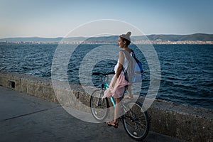 Young stylish woman in dress and with backpack is riding bicycle on promenade